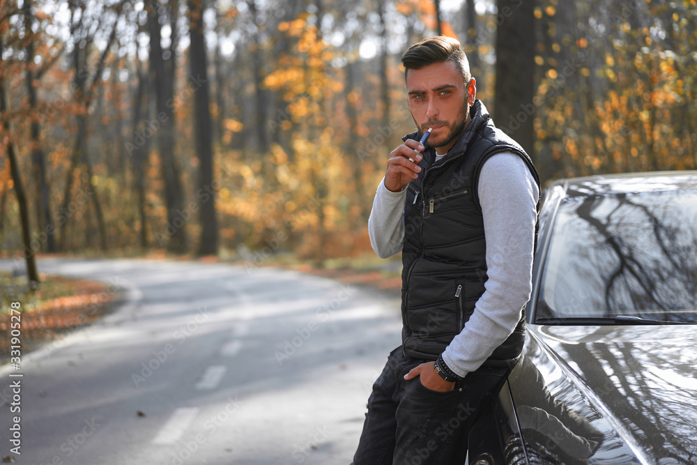 A handsome young Caucasian man wearing style black vest standing near black car on nature. A handsome young Caucasian man wearing style black vest standing near black car on nature.