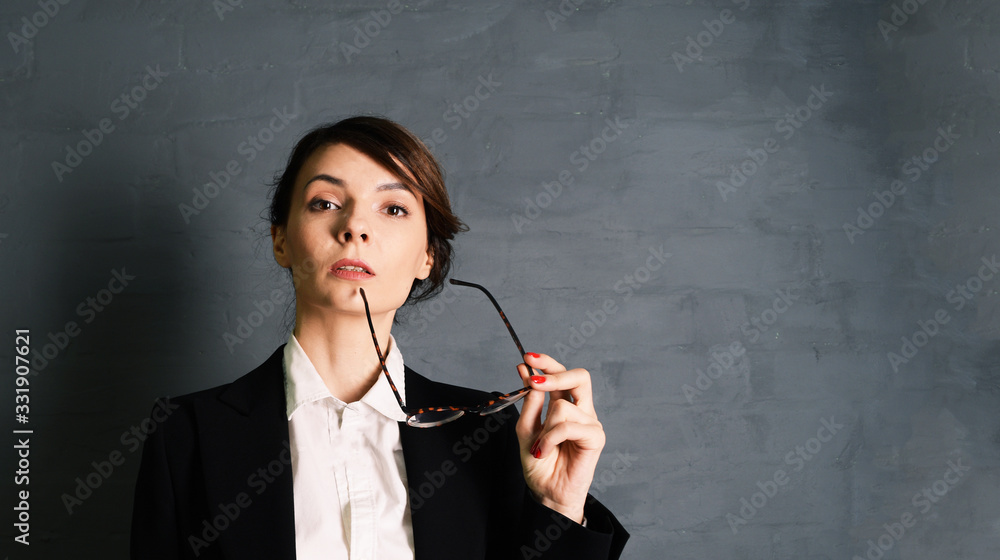 Young woman brunette teacher in a black jacket on a gray background holds glasses