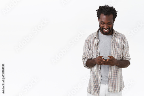 Modern young happy african-american bearded man in casual plain shirt, holding mobile phone, messaging via smartphone or use app to watch funny videos, smiling as look display, white background photo