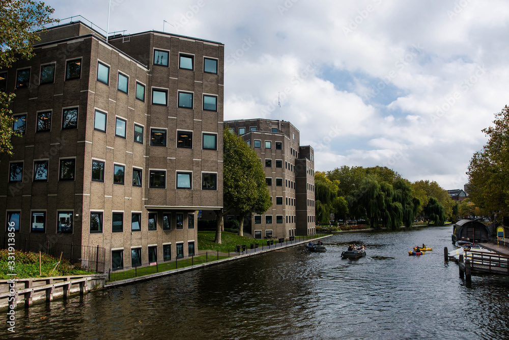 Travel to Amsterdam. Canal with boats. Modern exterior of new building. Green trees.