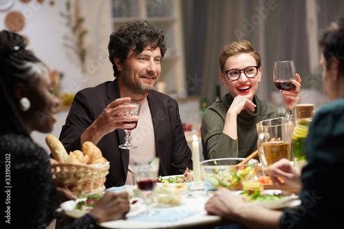 Young smiling couple drinking wine at the table and enjoying the company of their friends during holiday dinner at home