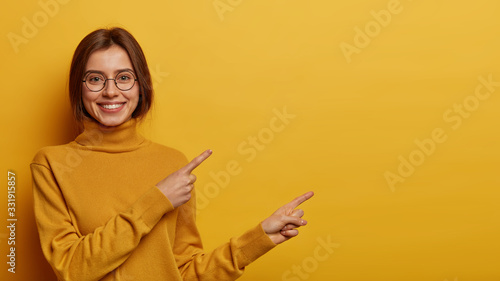 Horizontal shot of happy European woman with happy smile points with index fingers on empty space, shows place for your advertisement or promotional information, wears glasses and turtleneck © wayhome.studio 