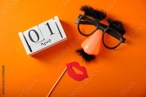 Wooden calendar with the date of April 1, on an orange background, false glasses, nose and mustache