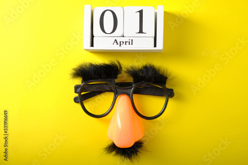 Wooden calendar with the date of April 1, on a yellow background, false glasses, nose and mustache
