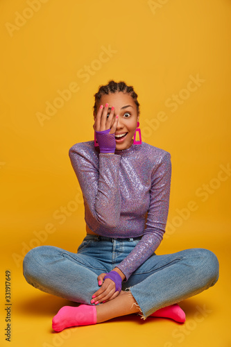 Vertical shot of glad beautiful ethnic woman keeps palm on face, has surprised happy expression, stares positively at camera, wears glittering purple jumper and jeans, isolated on yellow background © wayhome.studio 