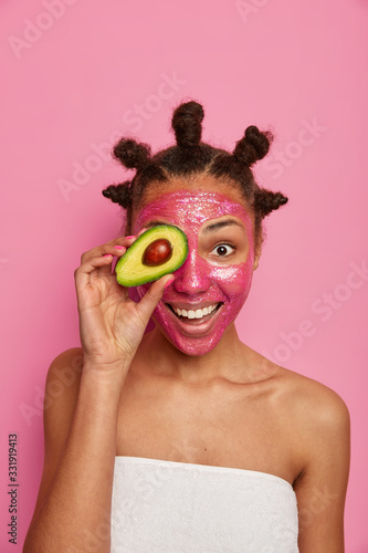 Fototapeta Naklejka Na Ścianę i Meble -  Positive young female model applies beauty face mask on face, smiles broadly, covers eye with half of avocado, has bare shoulders, wrapped in bath towel. People, wellness and cosmetology concept