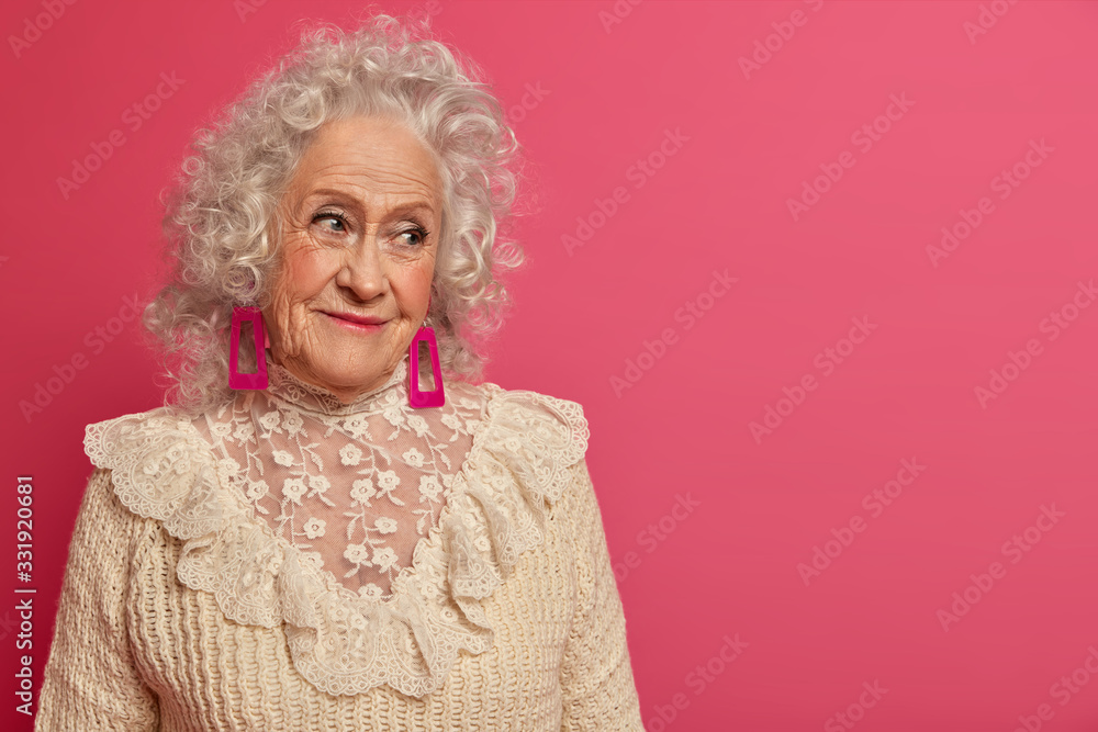 People, aging and cosmetology concept. Wrinkled grey haird senior woman gazes delighted aside, wears brown sweater, remembers nice memory, poses against pink background, empty space for your text