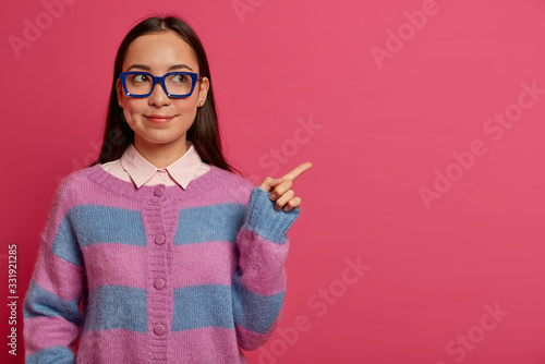 Beautiful clever female student in big glasses and jumper, introduces product promo, recommends click on link, shows item on pink background, presents new app for studying, shows best price.
