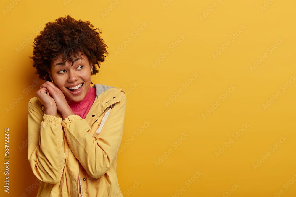 Beautiful cheering woman with overjoyed expression, keeps hands pressed together near face, looks aside right, dressed in casual anorak, poses against yellow background, copy space for promo Beautiful cheering woman with overjoyed expression, keeps hands pressed together near face, looks aside right, dressed in casual anorak, poses against yellow background, copy space for promo