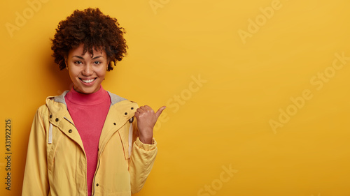 Studio shot of smiling woman has Afro hairstyle, wears yellow anorak, points thumb aside, chooses product, satisfied with good choice, recommends something to buy in store. Customes and sales