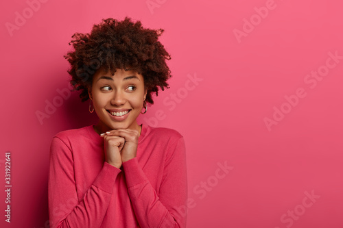 Portrait of positive dark skinned young woman keeps hands under chin  smiles toothily and looks aside  has pleased expression  wears crimson jumper  sees something appealing  creats plan in mind