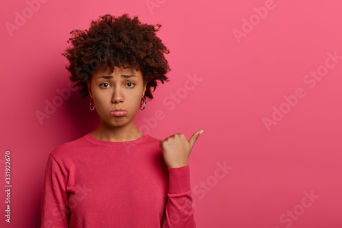 Gloomy sullen Afro American woman sulks with sad grimace, points thumb aside, begs to buy something, expresses regret emotions, doesnt have something she desires, gestures against crimson background