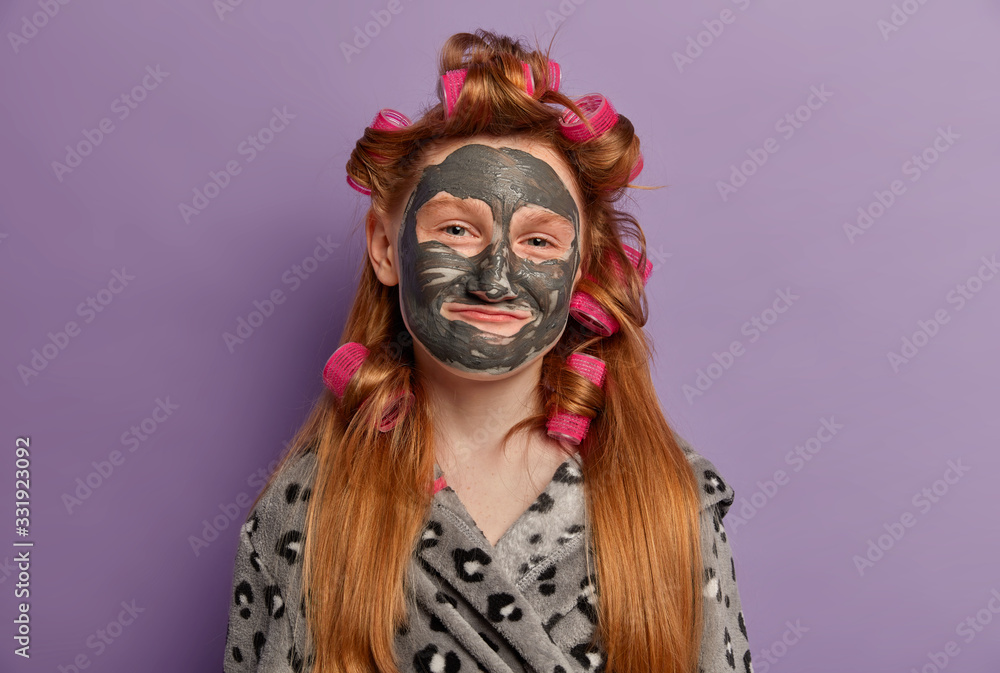 Little girl enjoys clay facial mask, has healthy glow perfect skin, wears hair curlers, looks happily at camera, has spa beauty treatments, spends free time at home, isolated on purple background