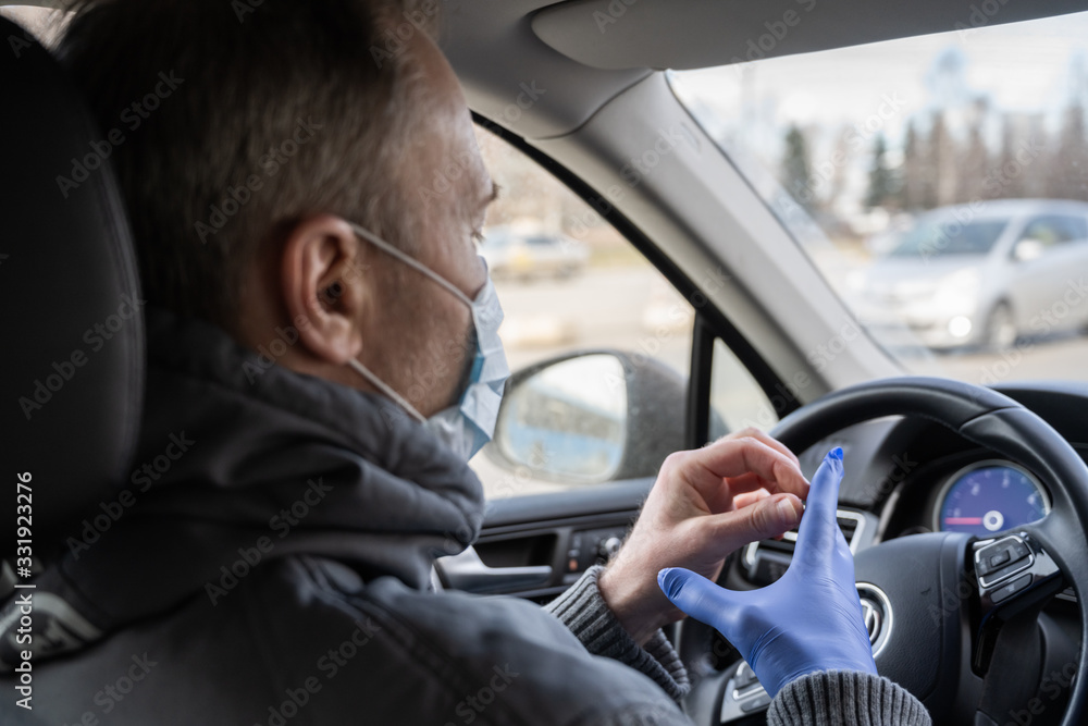 Man in the medical mask puts on rubber gloves for protect himself from bacteria and virus while driving a car. Coronavirus. Pandemic