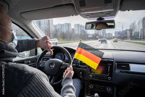 Middle aged man in the medical mask showing thumbs down with Germany flag driving a car. symbol of adverse epidemiological situation in countries. Coronavirus. Pandemic