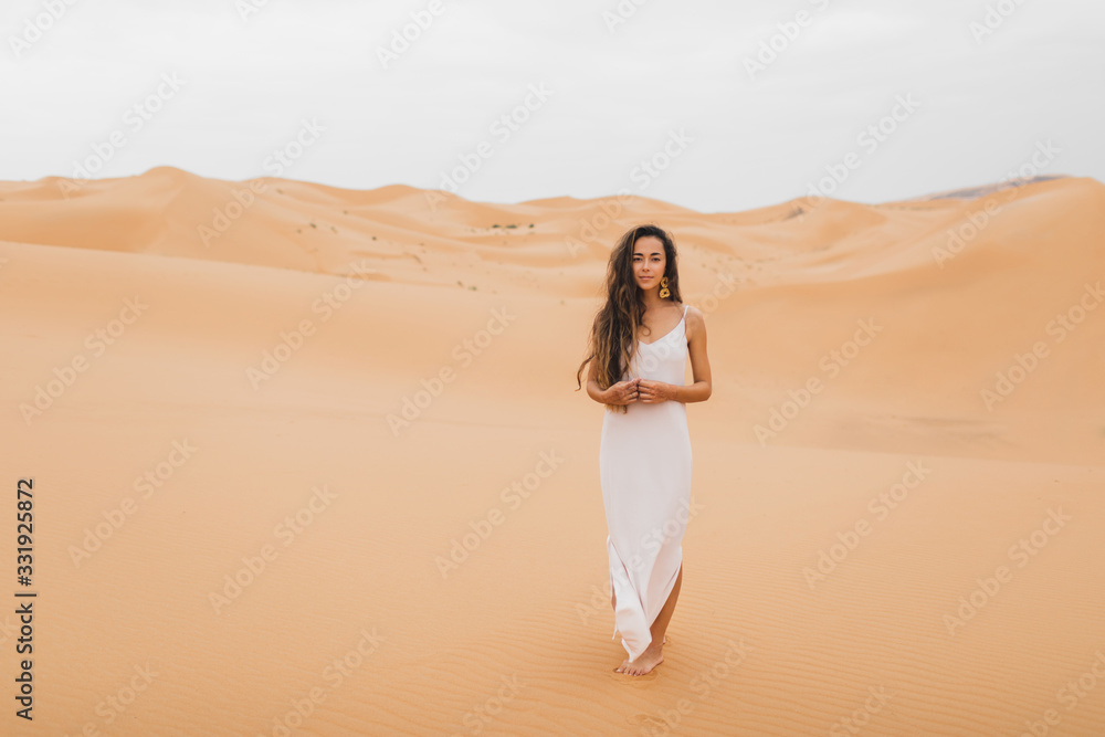 Pretty portrait of young beautiful woman in sand dunes of moroccan Sahara desert. Brunette with long hair, eastern appearance. Bride in white silk dress. Pretty portrait of young beautiful woman in sand dunes of moroccan Sahara desert. Brunette with long hair, eastern appearance. Bride in white silk dress.