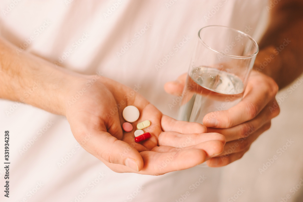 Drug addiction of young man. Sick patient holding oral medicaments and glass of water in hand. Man taking different pills. Treatment of coronavirus COVID-19 symptoms. Medicines or vitamins in hand Drug addiction of young man. Sick patient holding oral medicaments and glass of water in hand. Man taking different pills. Treatment of coronavirus COVID-19 symptoms. Medicines or vitamins in hand
