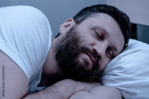 A lonely bearded man sleeps at night in bed with his arms folded under his head. Healthy sleep, pajamas, white bedding.