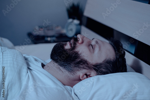 A lonely bearded man sleeps at night with his mouth open, snoring. Apnea syndrome, bedroom, pajamas, white bedding.