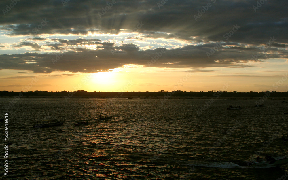 Pôr-do-sol em orla de rio com barcos em contraluz navegando e floresta na outra margem