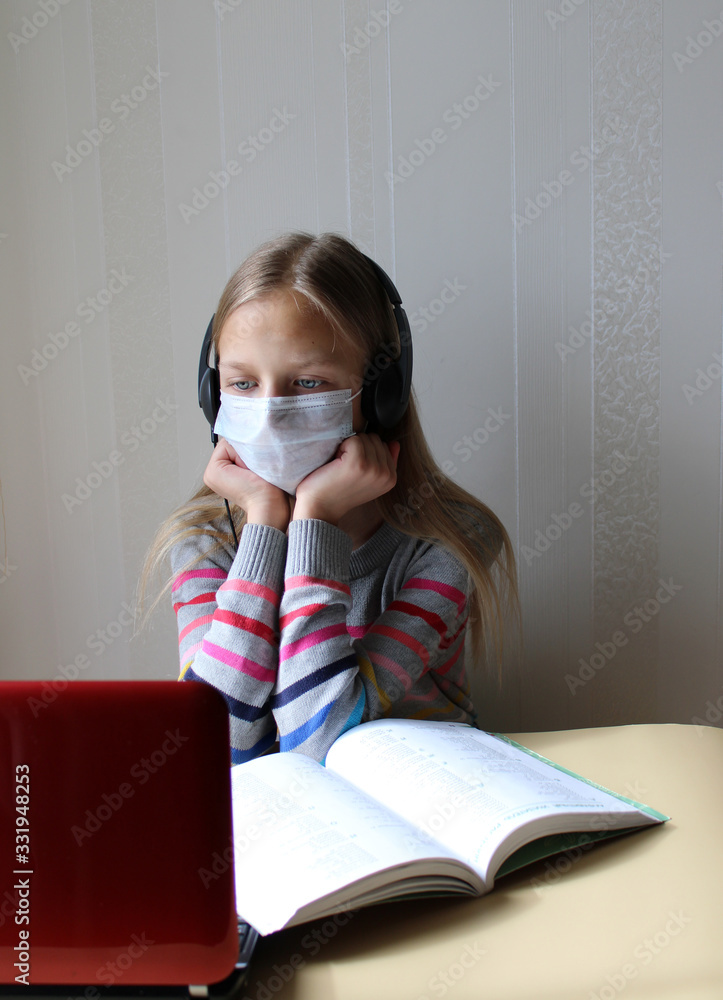 Blonde girl in a medical mask with headphones sits at a table with a computer and a book. Home schooling. The school is closed on quarantine due to the pandemic of Covid-19. Distance learning Blonde girl in a medical mask with headphones sits at a table with a computer and a book. Home schooling. The school is closed on quarantine due to the pandemic of Covid-19. Distance learning
