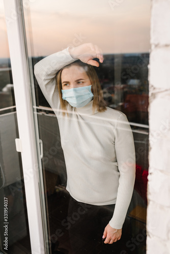 Sick woman wearing protection face mask looking through the window. Patient isolated in house to prevent infection. Quarantine room. Coronavirus.