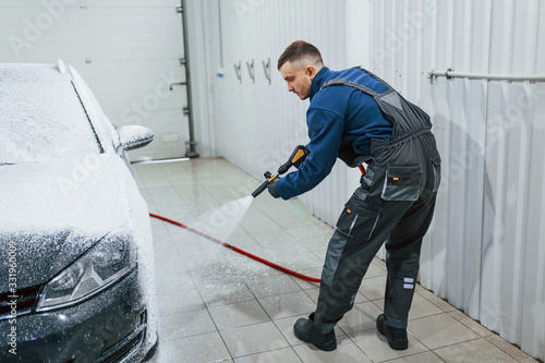 Male worker in uniform washing new modern car that covered with soap. Conception of service