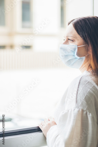 Young brown-haired woman, protected with a medical mask, looking from a window outside, COVID-19, coronavirus, selective focus