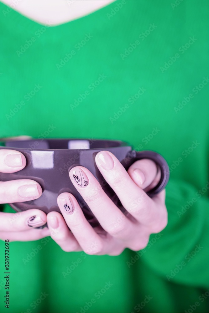 Fototapeta premium Beautiful female hands holding black cup on green background. Manicure with pink color nail polish, black floral pattern
