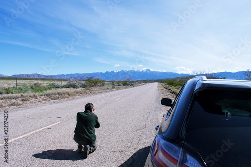 Photographer, man, kneeling on road taking photo of mountains in the distance with car on side of the road. 