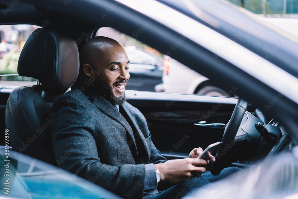 Happy adult ethnic employee using smartphone in car