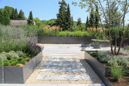 Moderne Garten- und Terrassengestaltung im Materialmix:Terrassen aus Steinplatten und Schotter sowie Metall Pflanzgefäße mit Gräsern, Bäumen und blühenden Kübel Pflanzen	