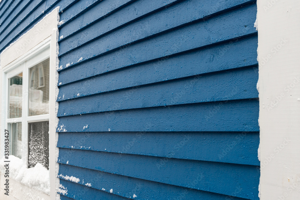 A royal blue wooden clapboard exterior wall of a vintage house. There's a white wooden four pane window and white edge trim on the structure. A royal blue wooden clapboard exterior wall of a vintage house. There's a white wooden four pane window and white edge trim on the structure.