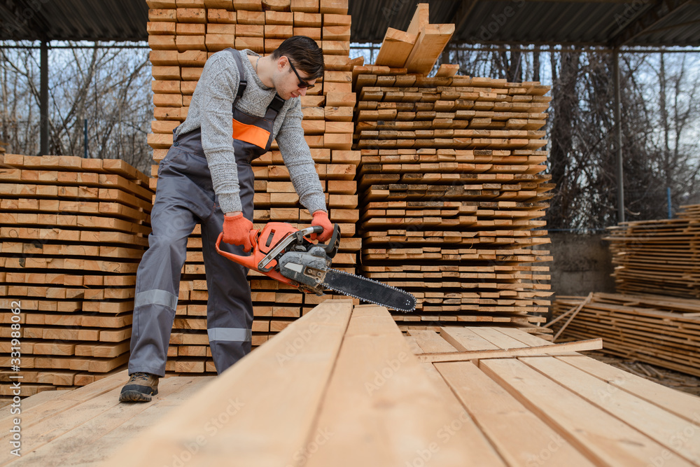 A portrait of young man carpenter at the wooden warehouse hold in his hands a chainsaw A portrait of young man carpenter at the wooden warehouse hold in his hands a chainsaw