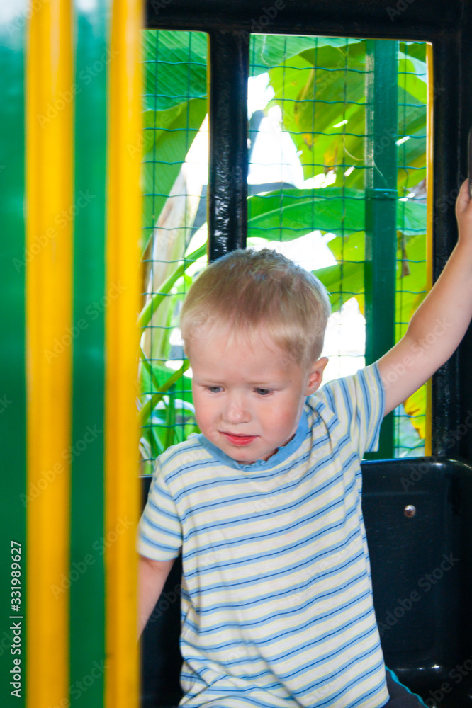Fototapeta premium A three-year-old little fair-haired boy rides in a yellow-green children's train in an amusement park in Riviera, Sochi, facing the viewer.