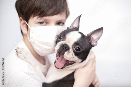 Friends-a boy in a medical mask and a Boston Terrier dog with a happy smile hug merrily. The concept of quarantine, virus, and isolation. © leksann