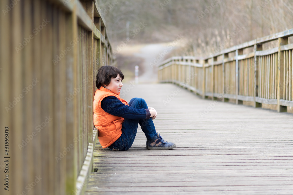 Pensive handsome boy sits on a wooden bridge.