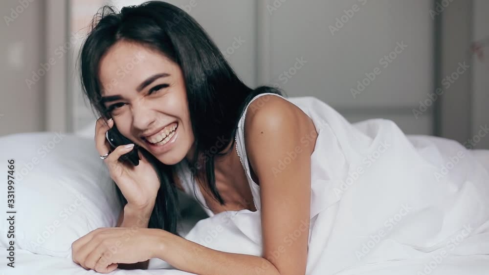 Beautiful young brunette woman smiling lying talking smart mobile phone relax in the bedroom Beautiful young brunette woman smiling lying talking smart mobile phone relax in the bedroom
