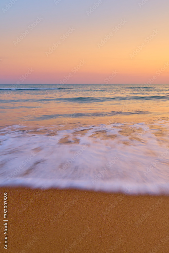 Colorful sunset at the tropical beach, sun behind clouds reflects on water and waves with foam hitting sand.