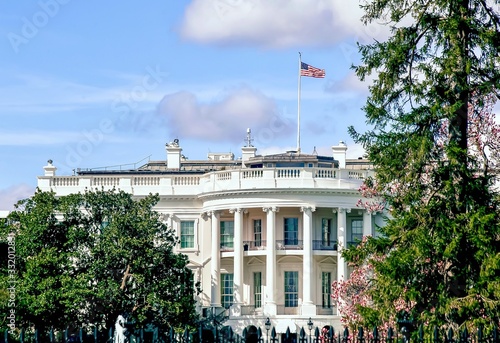 The United States White House on a beautiful sunny afternoon with an American flag on the roof with bright blue sunny sky and puffy white clouds.