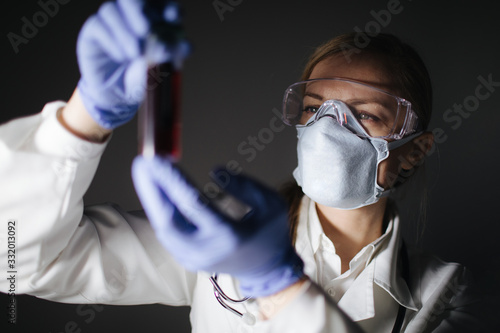 Concentrated female doctor in protective mask, eyewear and gloves holding glass bottle with blood sample for testing in laboratory. Dark studio background.
