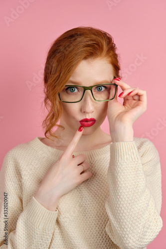 Red-haired girl with red lipstick and manicure adjusts her glasses and looks in the frame, putting her finger to her lips