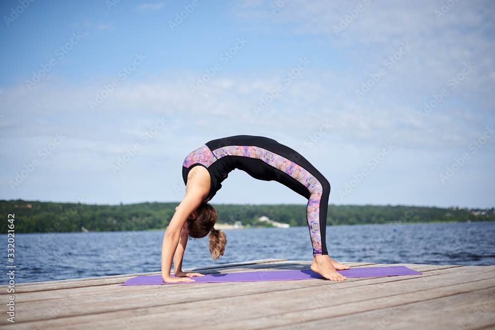 Young brunette woman with bare feet, wearing black and purple fitness outfit, doing gymnastic bridge pose on violet yoga mat on wooden pier in summer. Fit girl, stretching and doing yoga poses by lake