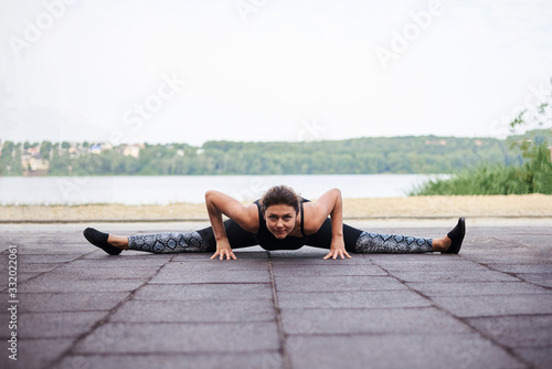Young brunette woman, wearing black fitness overall, doing stretching exercises by city lake in summer. Fit sportswoman on outside training. Healthy energetic active lifestyle concept.