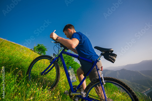 Perspective of a fit mountain biker pushing his bike uphill with amazing view on a forest, river and mountains in the background. Amazing green nature at sunset.
