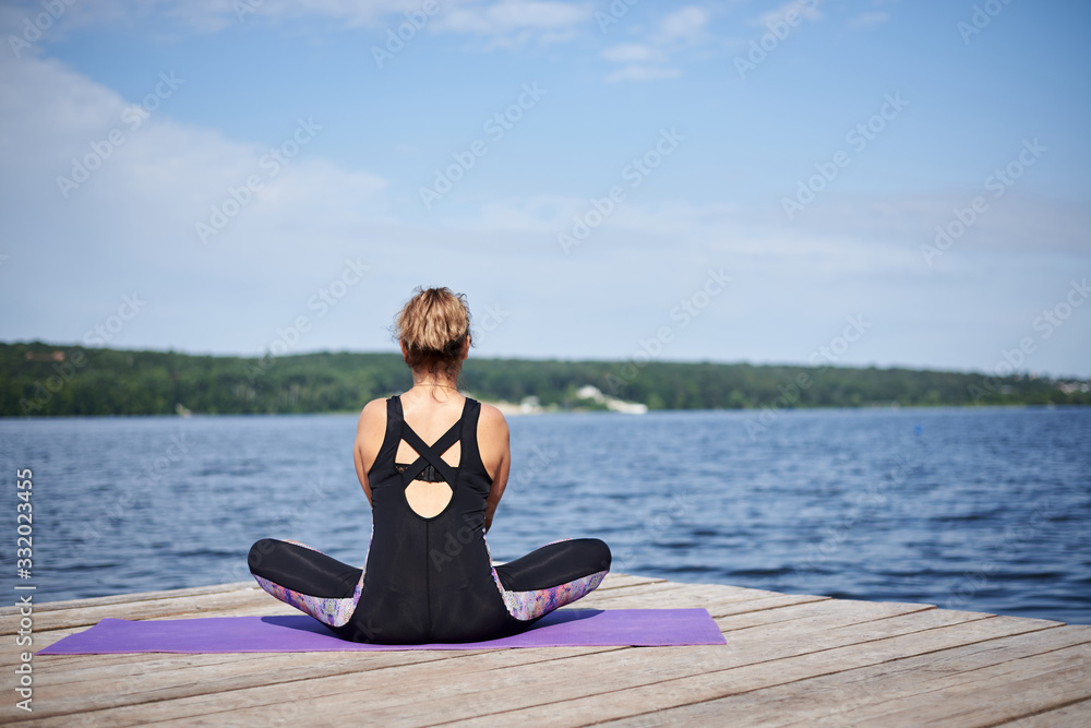 Young brunette woman, wearing black and purple fitness outfit, sitting on violet yoga mat outside on wooden pier in summer. Fit girl, doing yoga poses by lake, thinking, relaxing, meditating