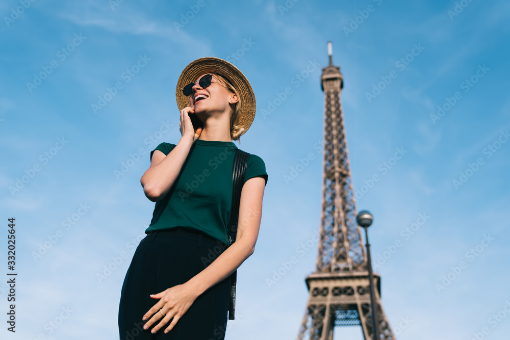 Pretty female tourist with sincerely smile on face enjoying international cellphone conversation via application, happy woman 20s using roaming for phoning rejoicing in Paris with Eiffel Tower