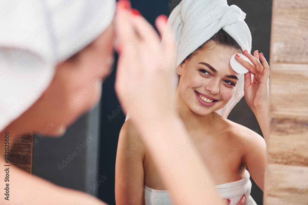 Beautiful young woman standing in bathroom, looking into the mirror and taking care of her face Beautiful young woman standing in bathroom, looking into the mirror and taking care of her face
