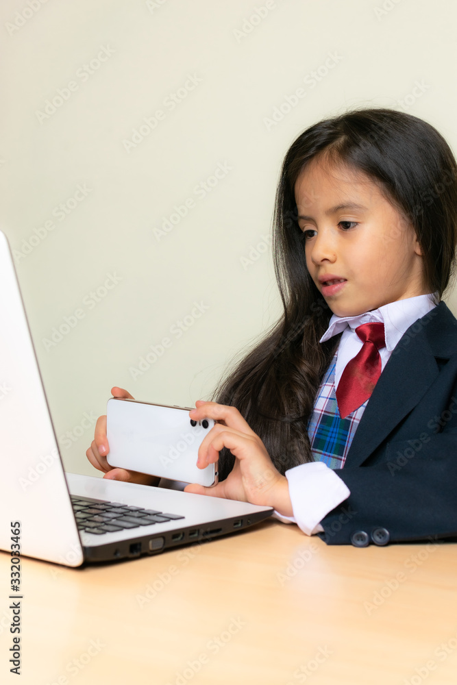 Colombian girl sitting at the desk in the classroom is doing homework. She takes a photo on the computer screen with her cell phone. Colombian girl sitting at the desk in the classroom is doing homework. She takes a photo on the computer screen with her cell phone.