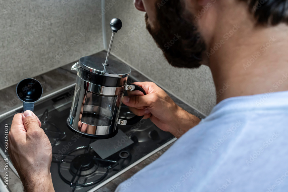 Close up of a person with brown hair's hands fixing coffee in a black and stainless steal metal french press over a small stove covered in glass and grey counter top inside one early morning.